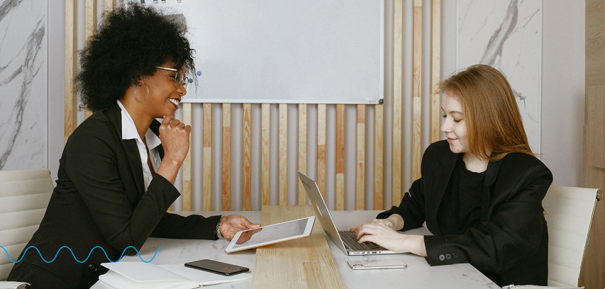 Two business people sit at a modern office table and converse while using electronic devices